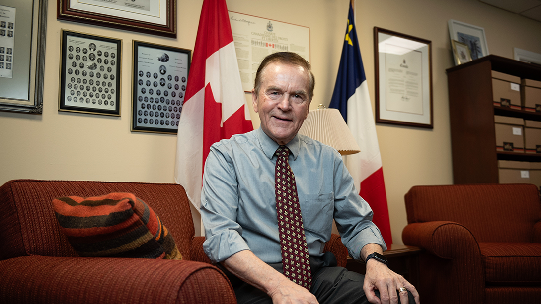 Senator Percy Mockler sits on a chair in his office with the Canadian and Acadian flags behind him.