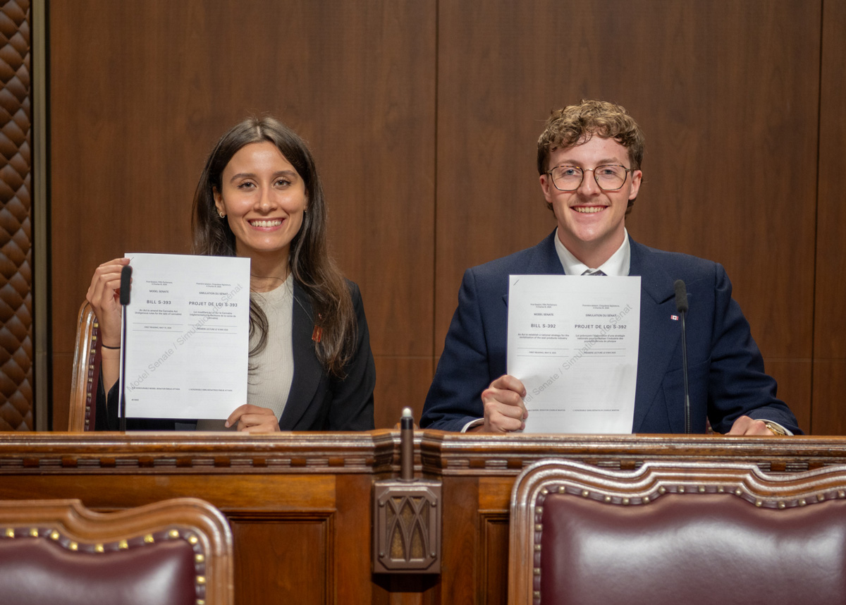 A young woman and a young man, sitting in the Senate Chamber, proudly hold a copy of a bill.