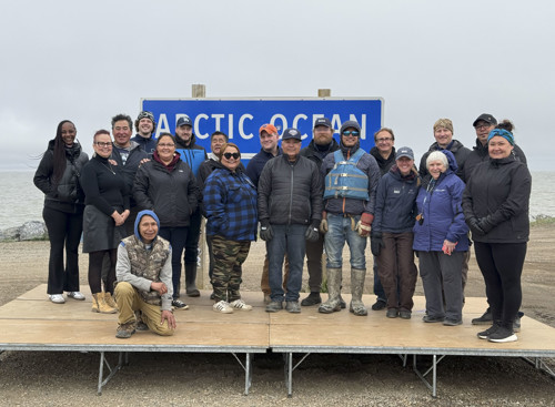 A group of people pose on a wooden platform in front of an 'Arctic Ocean' sign, with the ocean in the background. 