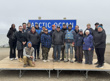 A group of people pose on a wooden platform in front of an 'Arctic Ocean' sign, with the ocean in the background. 