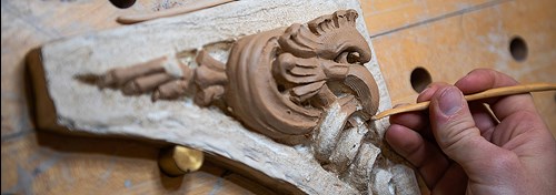 A closeup of a hand shaping a carving of a cockatrice using a clay-sculpting tool.