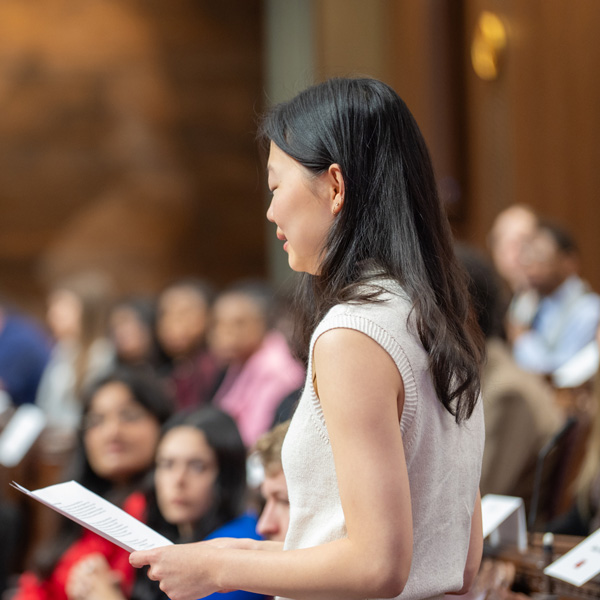 A young woman stands holding a sheet of paper, surrounded by a group of people sitting in the background.