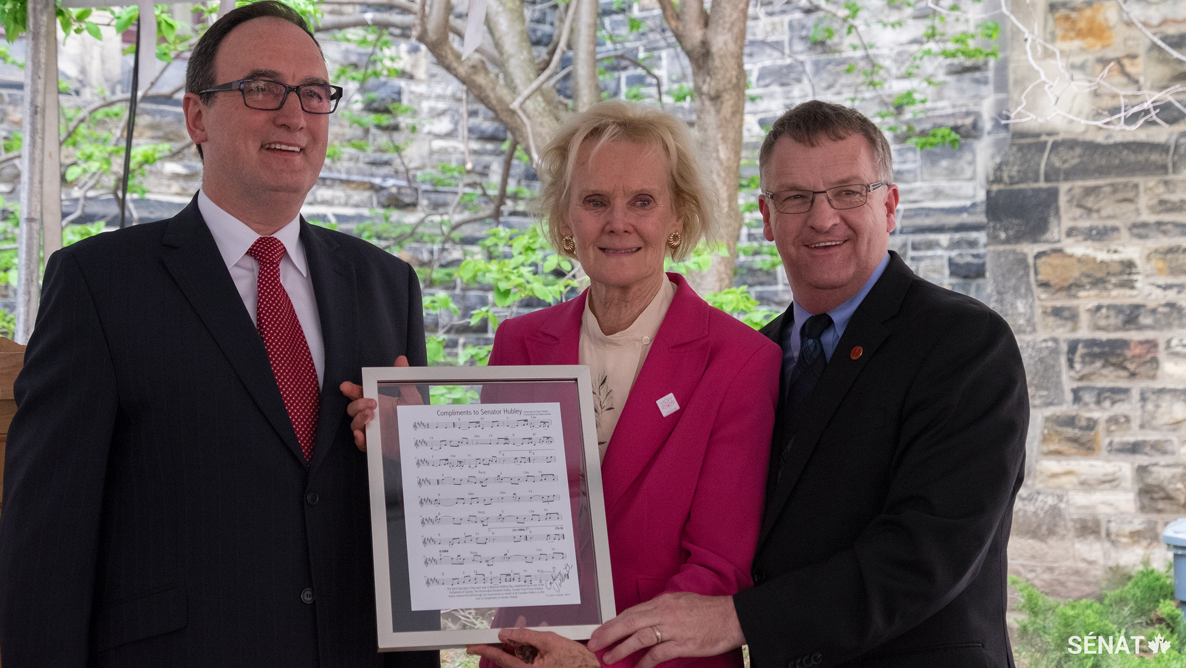 Graham Sheppard, président de l’Association canadienne des grands maîtres violoneux (à gauche) est accompagné du Sénateur Fabian Manning (à droite) pour présenter à la sénatrice Hubley un exemplaire encadré de la feuille de musique d’une pièce créée en son honneur.