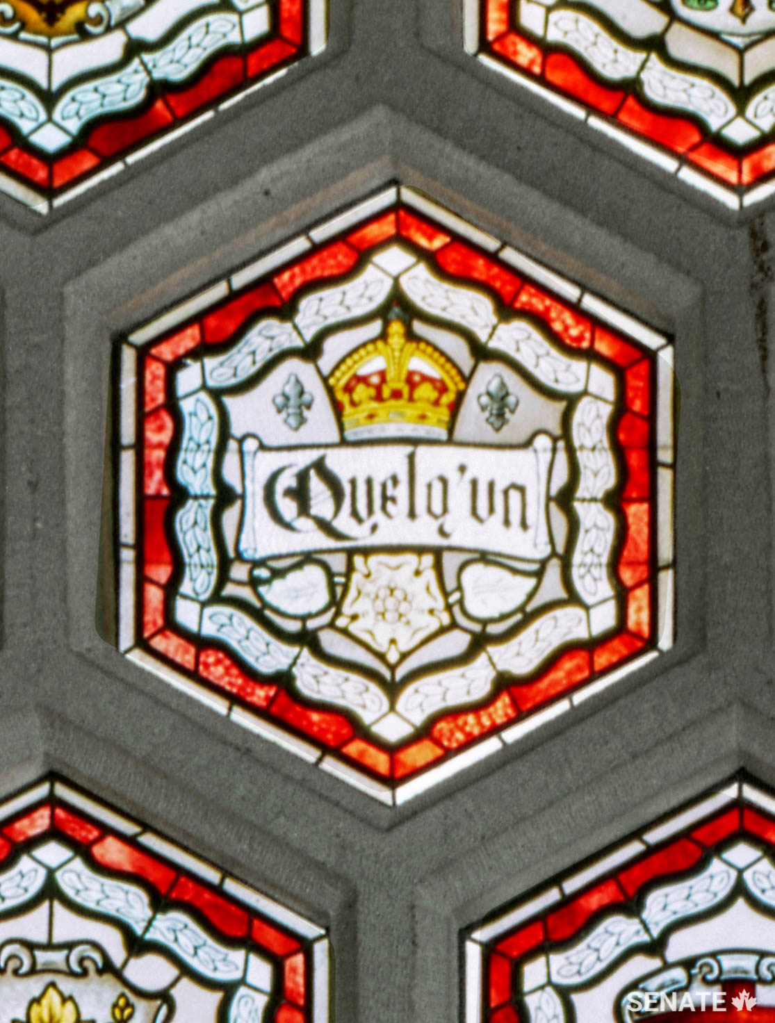 The foyer’s stained-glass ceiling incorporates the names of all the speakers of the Senate until 1920. A single pane with a cryptic message represents Senate speakers to come. It is not clear whether the misspelling (it should be <em>quelqu’un</em>) is intentional.