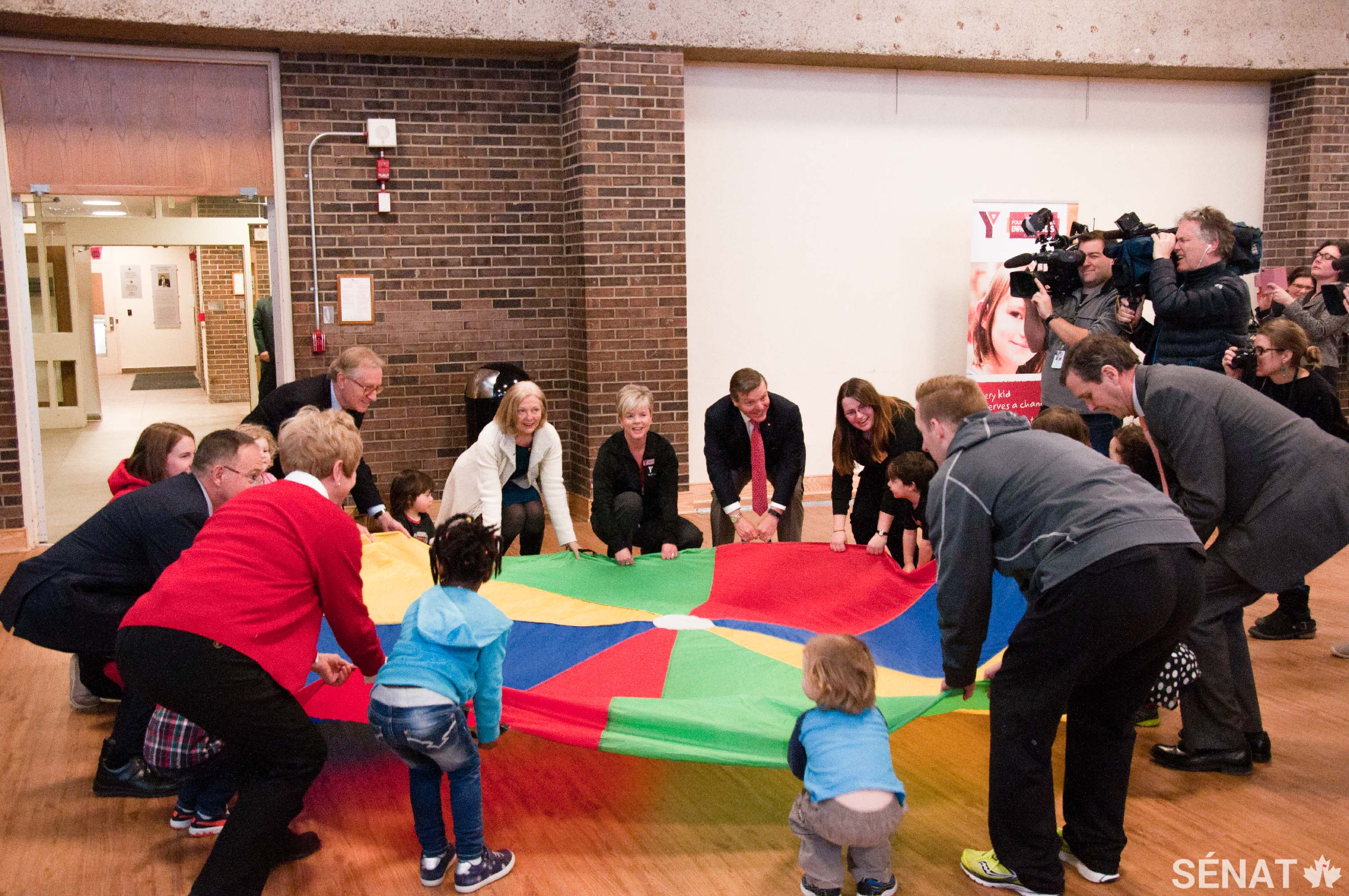 Des membres du Comité sénatorial des affaires sociales, des sciences et de la technologie participent à une activité de parachute avec des enfants à un YMCA d’Ottawa avant leur conférence de presse de mars 2016 qui visait à annoncer la publication de leur rapport sur l’obésité au Canada.