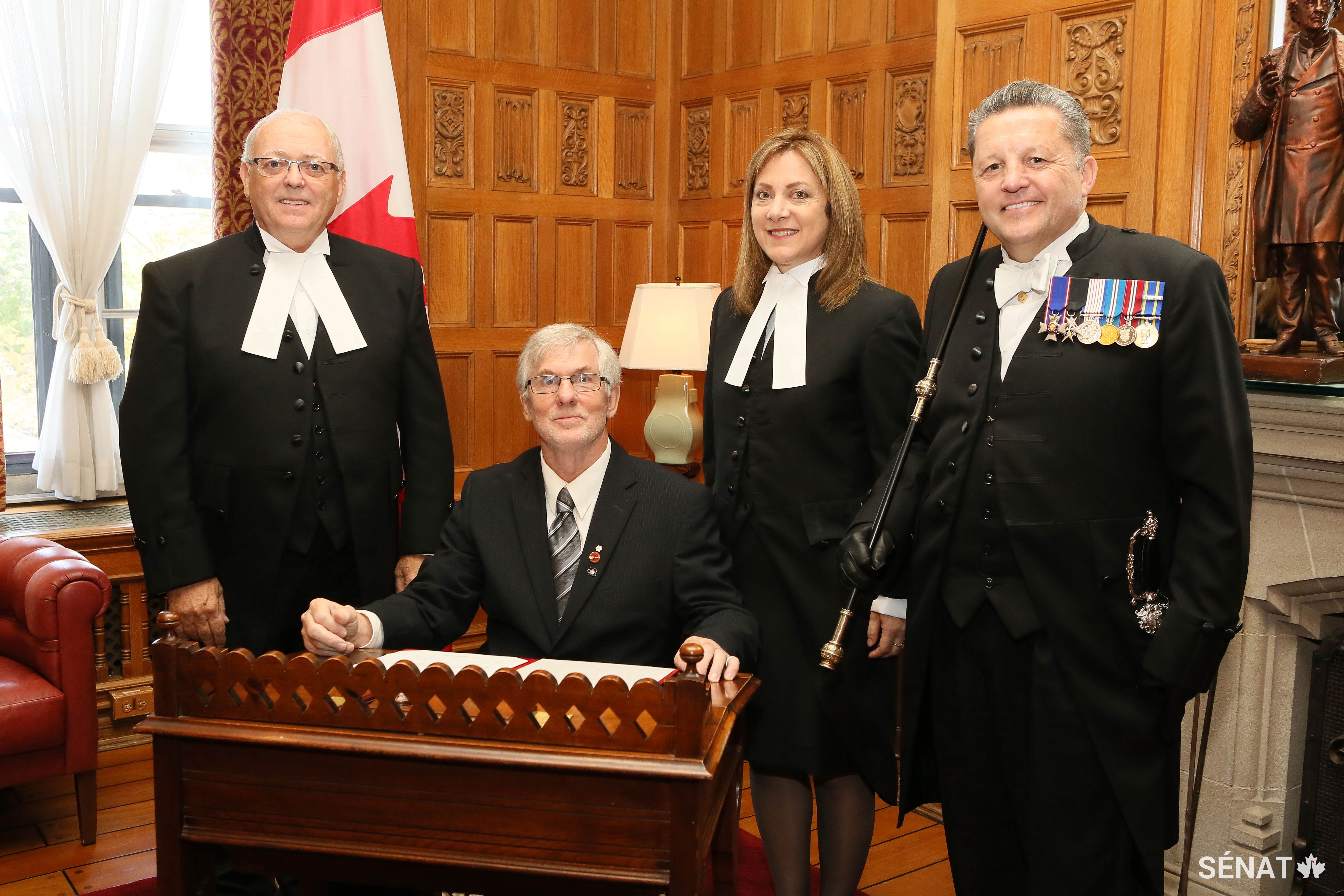 Le sénateur David Adams Richards (assis) se joint à la Chambre haute, entouré du <a href='https://sencanada.ca/fr/presidents/georgefurey/biographie/'>Président du Sénat, George J. Furey</a>, de la greffière du Sénat, Nicole Proulx, et de l’<a href='https://sencanada.ca/fr/a-propos/huissier-du-baton-noir/'>huissier du bâton noir, J. Greg Peters</a>.