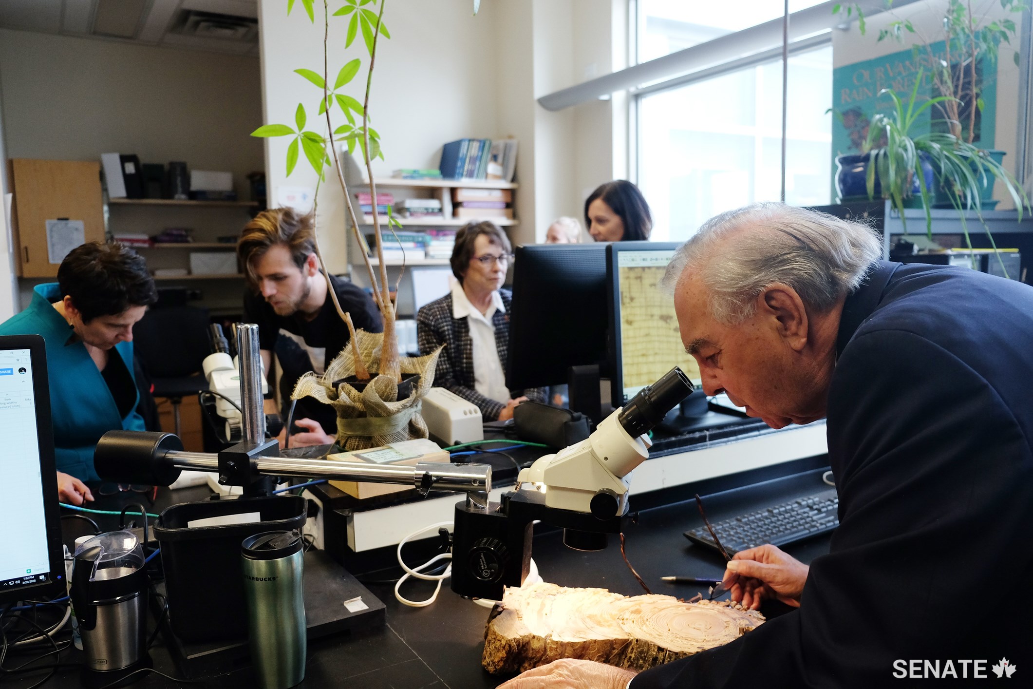 Senator Ghislain Maltais uses a microscope to get a closer look at a wood core sample while Senators Raymonde Gagné and Diane F. Griffin listen to students.