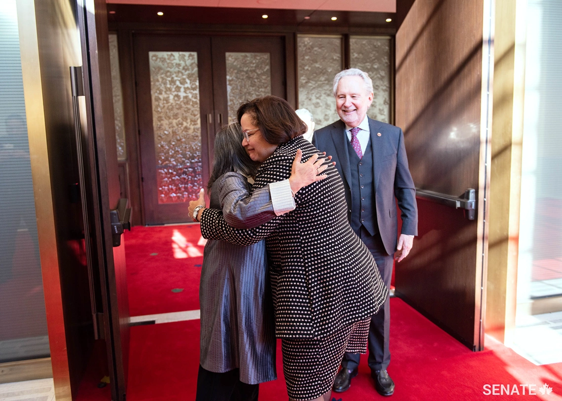 Senator Peter Harder smiles as Senator Ratna Omidvar hugs recently sworn-in Senator Rosemary Moodie on February 19, 2019.
