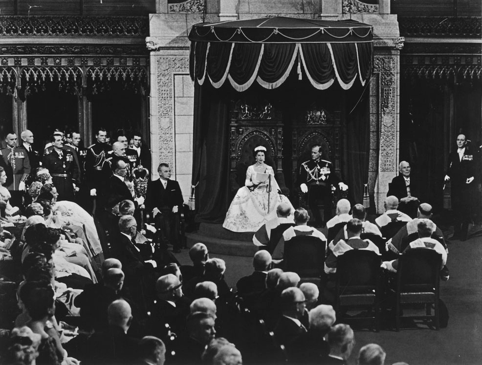 Dans la Chambre du Sénat avec le prince Philip à ses côtés, la reine Elizabeth II lit le discours du Trône lors de l’ouverture de la 23e législature du Canada, le 14 octobre 1957. (Bibliothèque et Archives Canada)