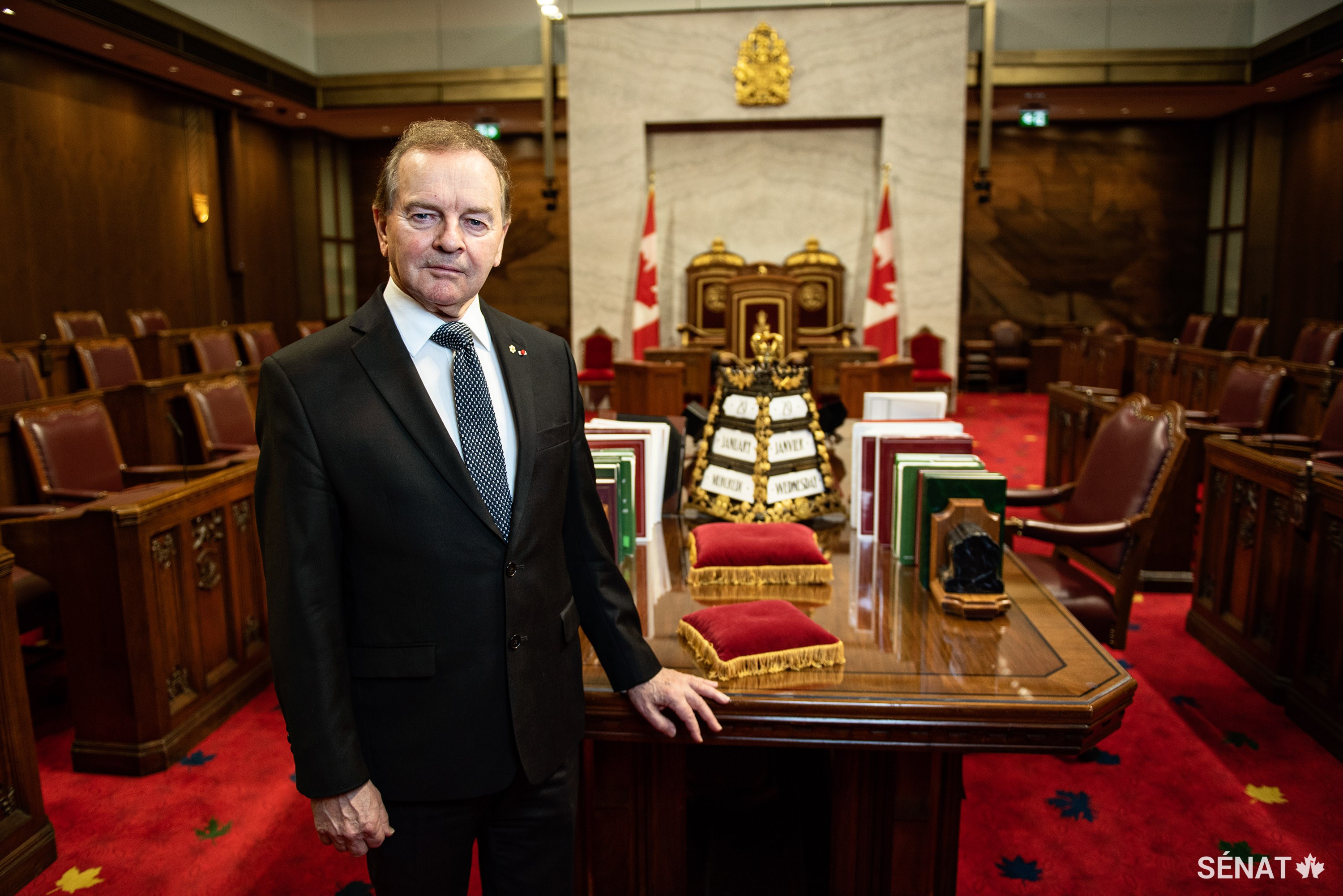 Le sénateur Serge Joyal pose à côté du calendrier qu'il a pris l’initiative de faire réaliser pour marquer le jubilé de diamant de Sa Majesté. Le calendrier posé sur le Bureau du greffier a été inauguré en 2013. Il a été financé par des dons privés de sénateurs anciens et actuels, de greffiers et de cadres supérieurs du Sénat. Réalisé en laiton et bronze, il est orné de symboles monarchiques et canadiens rehaussé de dorure.