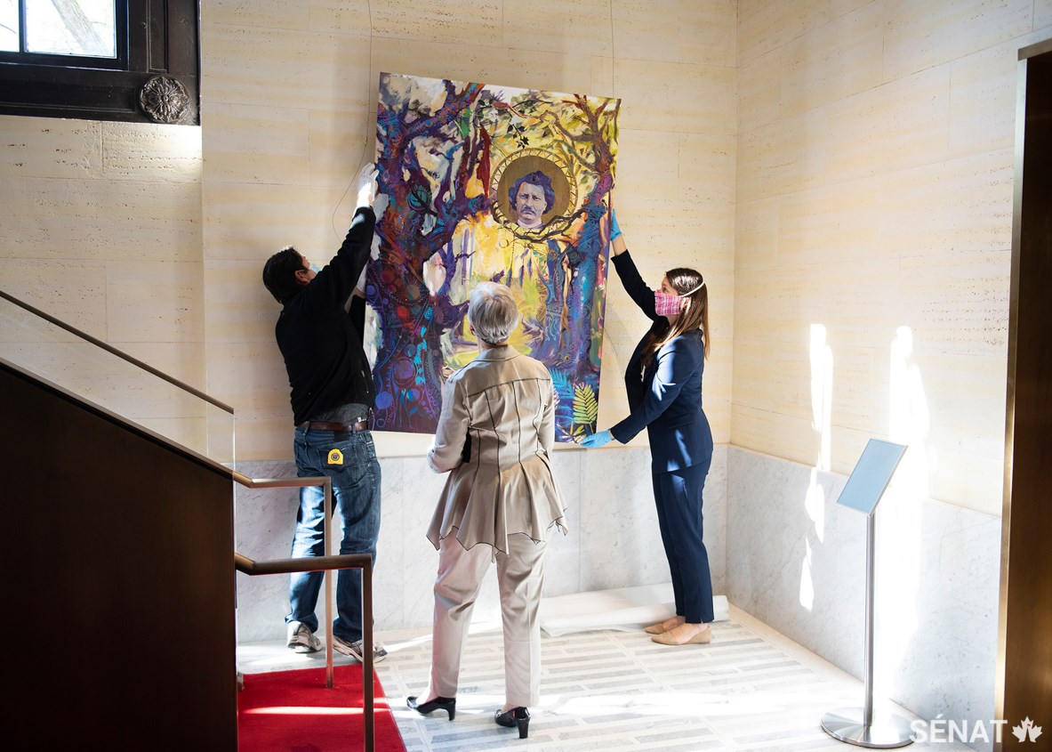 La sénatrice Patricia Bovey (au centre) et Tamara Dolan, la conservatrice du Sénat (à la droite), aident à installer Stolen Identities — un tableau de l’artiste nigérian-canadien Yisa Akinbolaji — à l’édifice du Sénat du Canada, le vendredi 18 septembre 2020.
