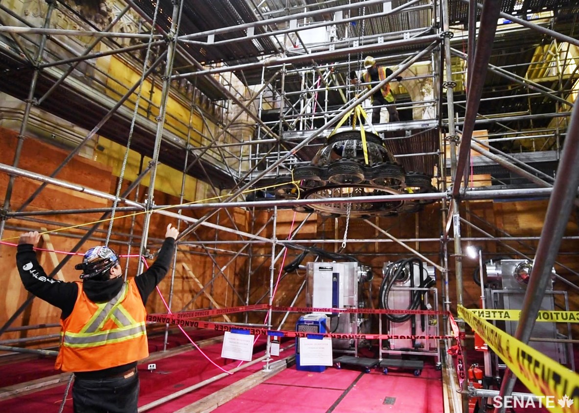 Workers slowly lower one of the stripped-down chandeliers using a heavy-duty chain pulley system. Structural nylon straps — akin to ultra-durable seatbelts — are wrapped around the strongest components of the chandelier and hooked to the pulley system.