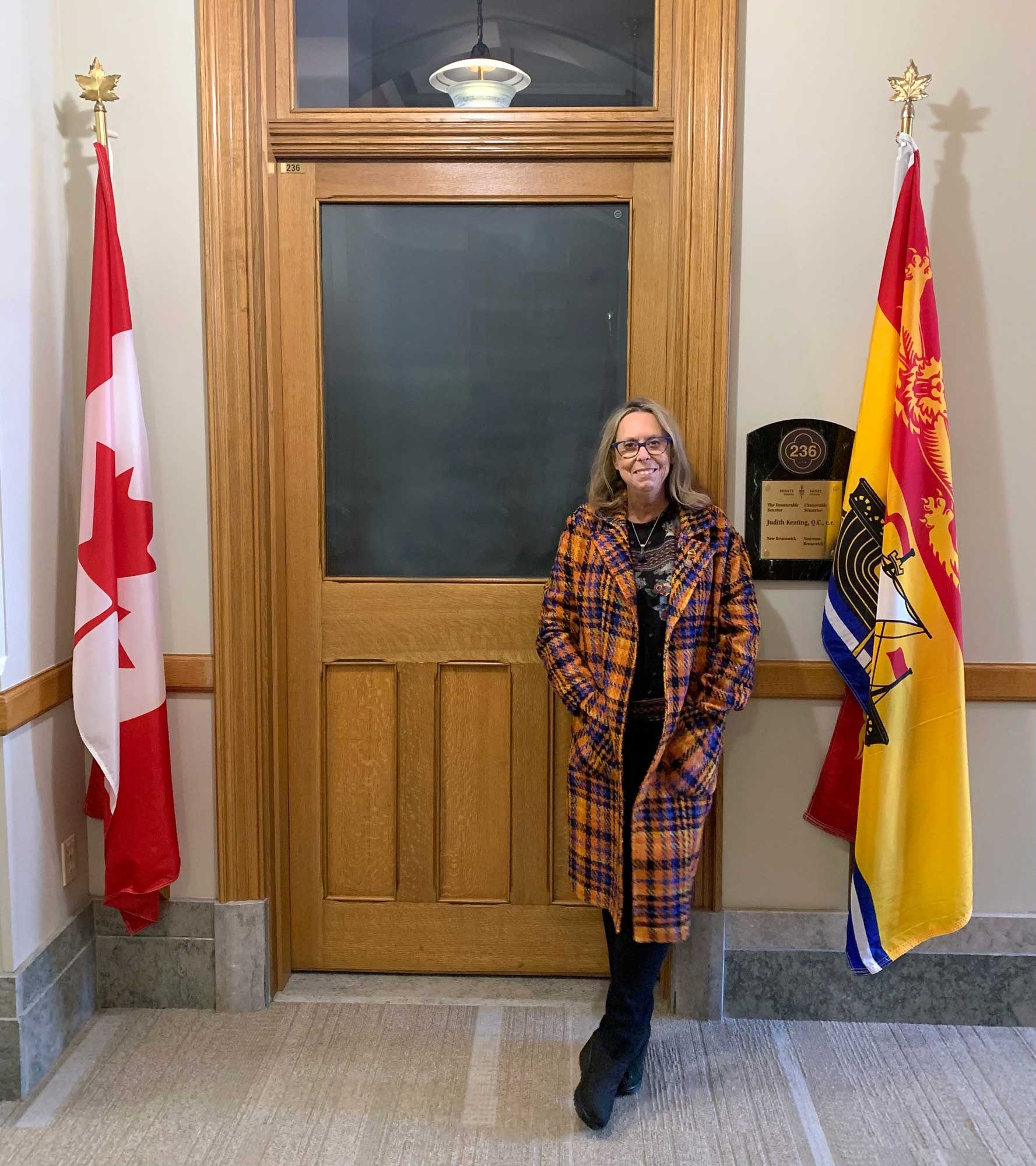 La sénatrice Keating devant la porte de son bureau au Sénat, flanquée du drapeau du Canada et de celui du Nouveau-Brunswick, la province qu’elle représentait à la Chambre haute. (Crédit photo : Bureau de la regrettée sénatrice Keating)