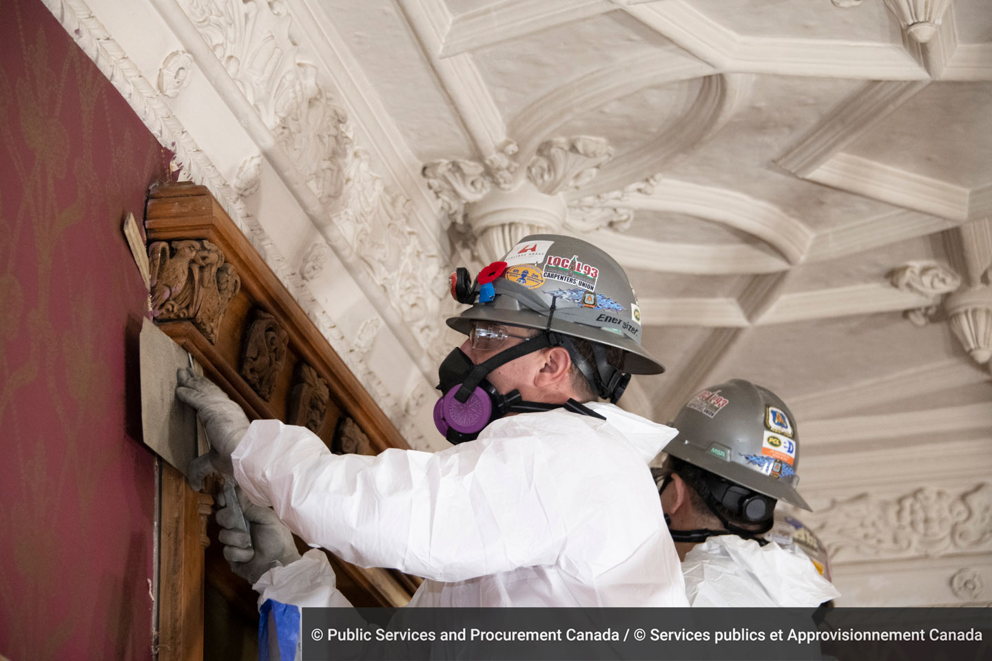 A worker from Heritage Grade holds a protective flat board against the wood frame at the top of the window surround as he slips a hacksaw blade underneath the wood to sever the nails.