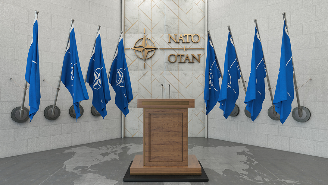 A wood podium with two microphones, in front of a wall with the NATO name and logo, flanked by four blue and white NATO flags on each side. 