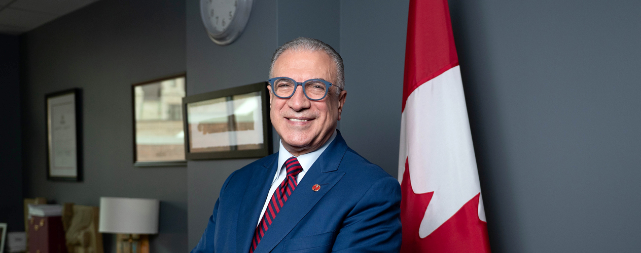 Un homme portant un costume, une cravate et des lunettes pose à côté d’un drapeau canadien.