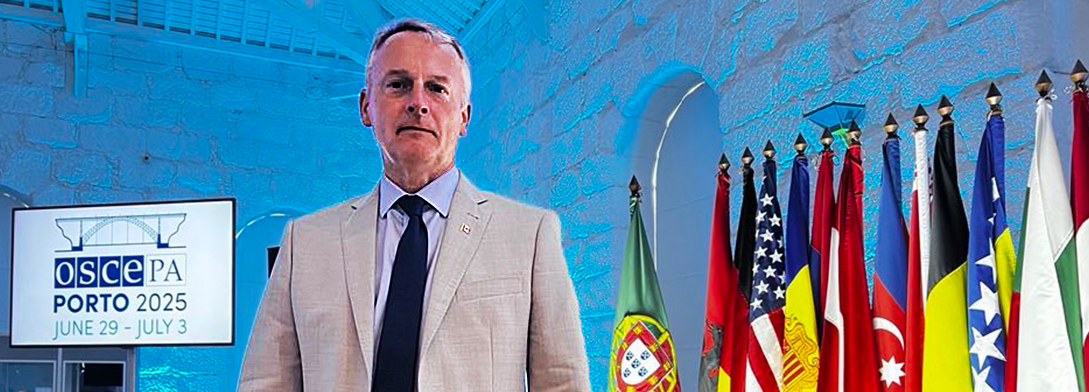 Senator David M. Wells stands in front of a row of international flags and a sign for the Organization of Security and Co-operation in Europe Parliamentary Assembly.