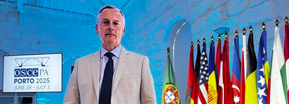 Senator David M. Wells stands in front of a row of international flags and a sign for the Organization of Security and Co-operation in Europe Parliamentary Assembly.