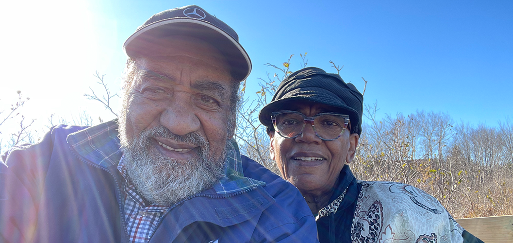 A Black man and a Black woman, both smiling and wearing hats, pictured on a sunny day.