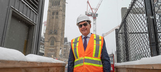 Un homme vêtu d’équipement de protection individuelle et de lunettes de soleil se tient debout sur un chantier de construction pendant une journée enneigée, avec la Tour de la Paix de l’édifice du Centre et une grue en arrière-plan.