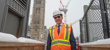 Un homme vêtu d’équipement de protection individuelle et de lunettes de soleil se tient debout sur un chantier de construction pendant une journée enneigée, avec la Tour de la Paix de l’édifice du Centre et une grue en arrière-plan.