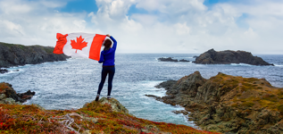 Une femme tenant le drapeau canadien, debout sur un rivage rocheux surplombant l’océan, sous un ciel bleu et nuageux.