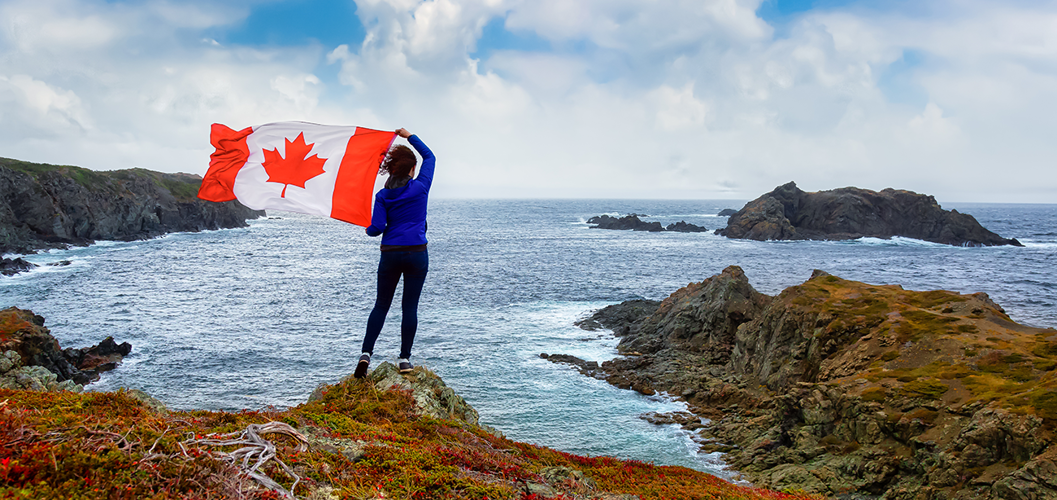 A woman holding the Canadian flag as she stands on a rocky shore overlooking the ocean, underneath a blue and cloudy sky.