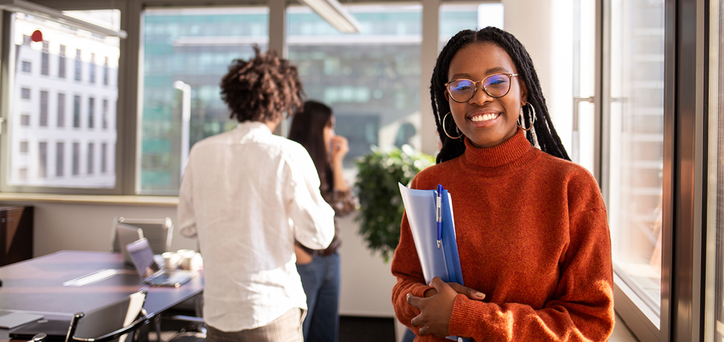 A young Black woman wearing glasses and an orange turtleneck holding a folder in an office.