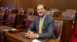 Senator Marc Gold, wearing a grey suit and yellow tie, sits at his desk in the Senate Chamber.