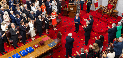 A woman standing in front of a throne is applauded by a crowd of people in the Senate Chamber.