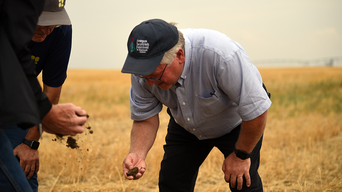 Senator Rob Black, wearing a ball cap, bends over in a farm field and inspects soil in his right hand.