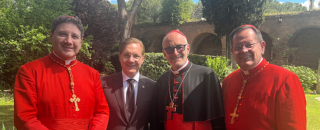 Four men pose in a garden; one wears a suit and the other three wear cassocks.