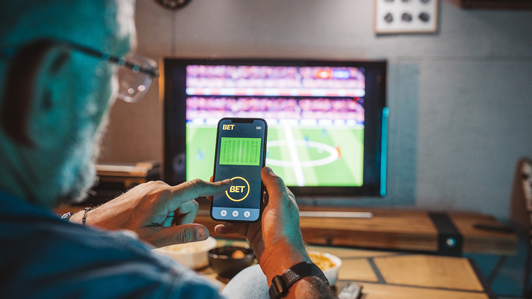 A man’s finger hovers over a button labelled “Bet” on a mobile sports gambling app, with a TV displaying a sports game in the background.
