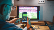 A man’s finger hovers over a button labelled “Bet” on a mobile sports gambling app, with a TV displaying a sports game in the background.