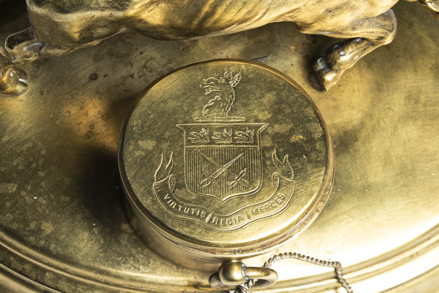 The lid of a cylindrical, brass inkpot, engraved with a crest and the Latin phrase “Virtutis regia merces,” which means “A palace the reward of bravery.”