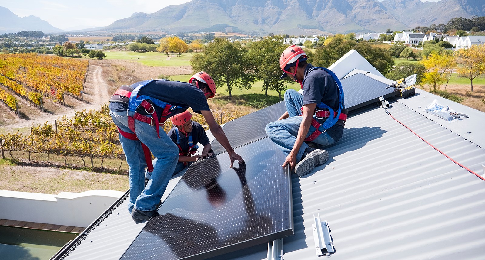 Three workers wearing protective gear install solar panels on a roof, with the landscape of Cape Town, South Africa behind them.
