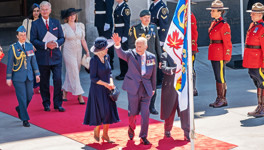 King Charles III waves from the red carpet in front of the Senate of Canada Building with the Personal Canadian flag of the monarch in the foreground.
