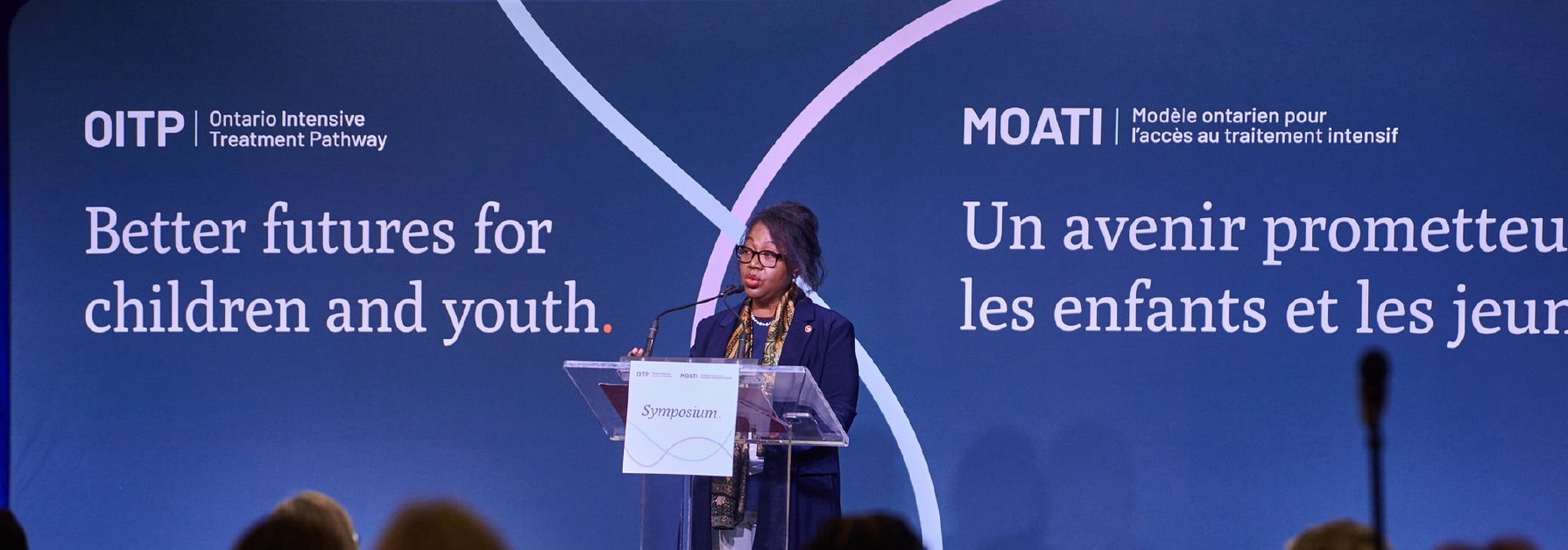 Une sénatrice s’exprime à un pupitre sur une scène lors d’une conférence, avec une grande bannière bleue derrière elle portant le message « Un avenir prometteur pour les enfants et les jeunes ».