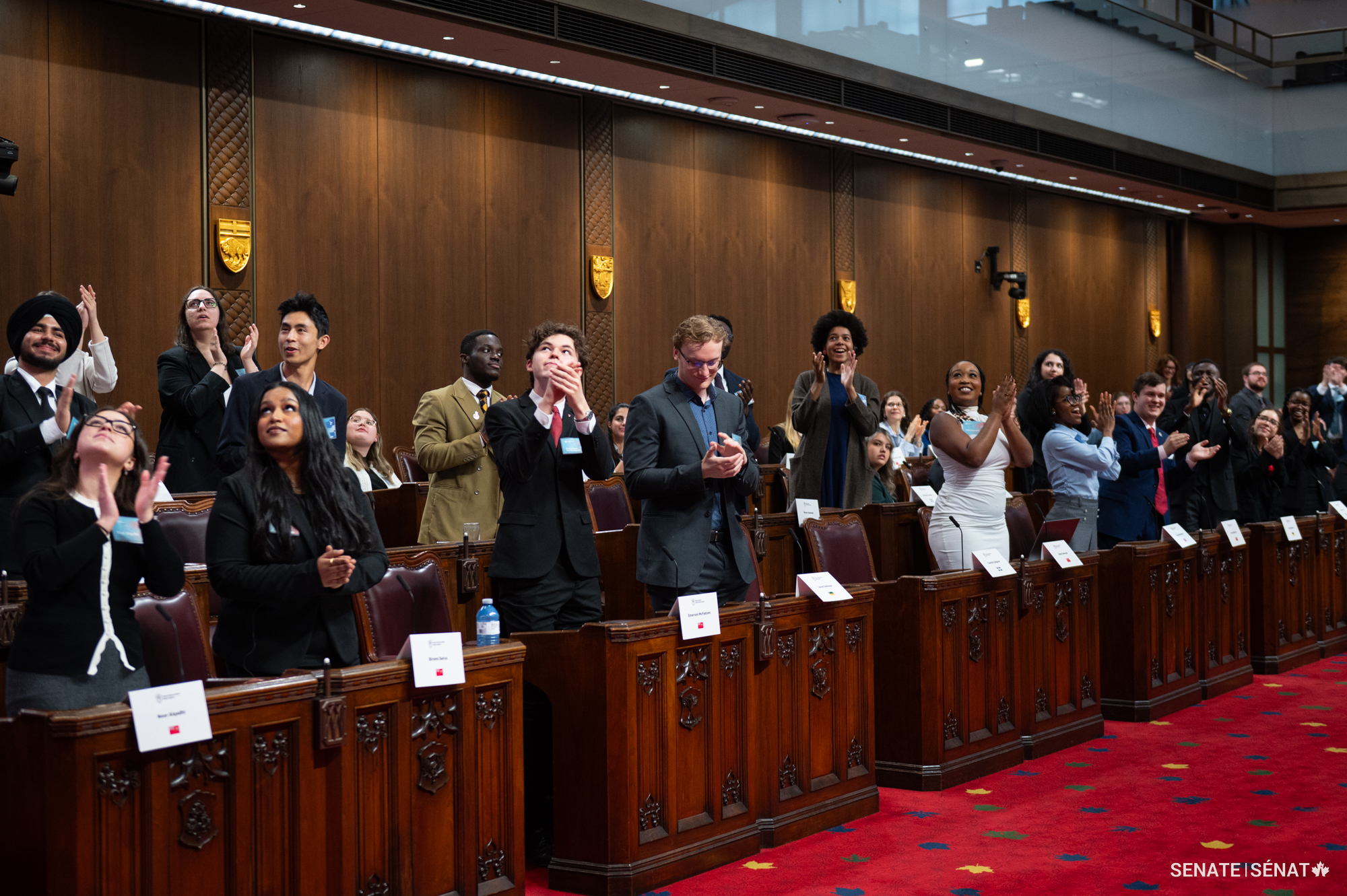 De jeunes adultes debout devant des pupitres en bois dans la Chambre du Sénat, applaudissant tout en levant les yeux.