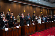 Young adults standing at wooden desks in the Senate Chamber clapping while looking upwards.