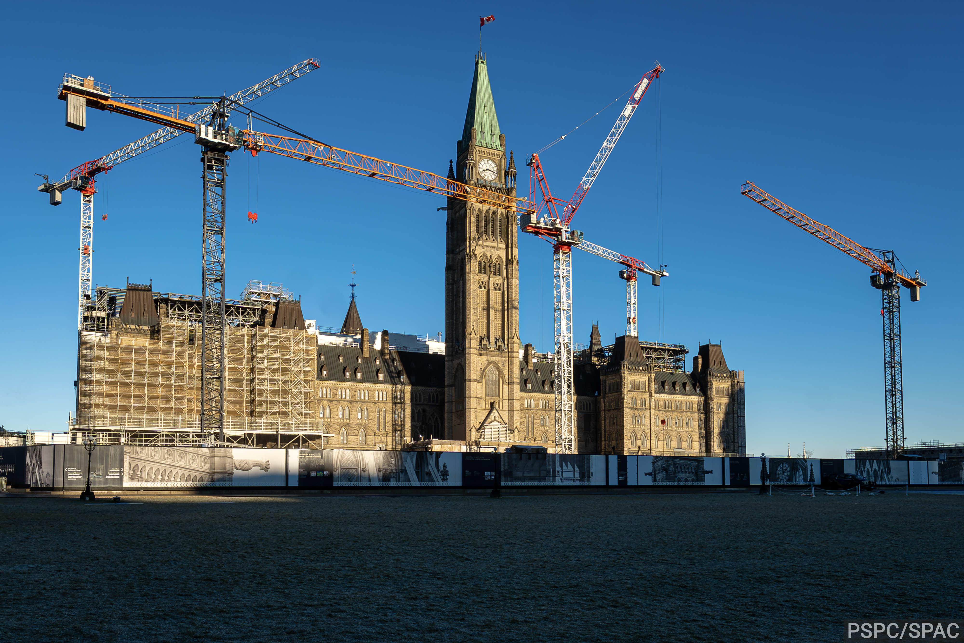 L’édifice du Centre sur la Colline du Parlement, baigné par la lumière de l’après-midi et entouré de cinq grues à tour et de panneaux de construction.
