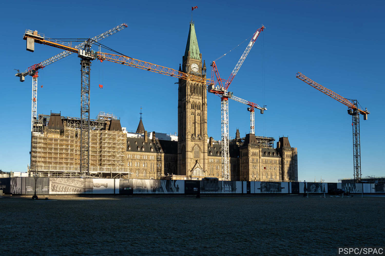 Centre Block on Parliament Hill, bathed in afternoon light and surrounded by five tower cranes and construction hoarding.