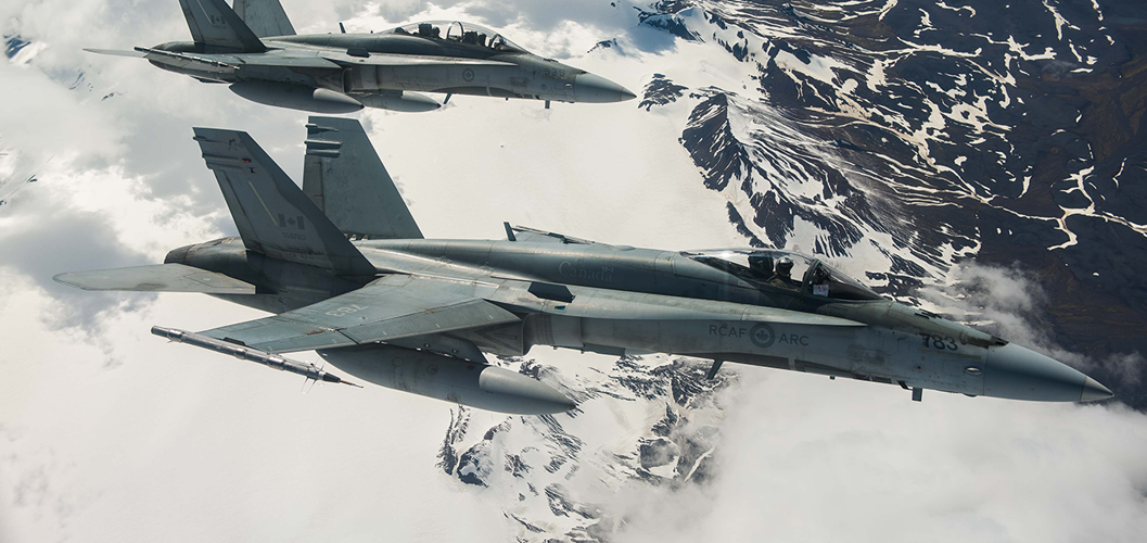 Deux avions de chasse gris de l’Aviation royale canadienne survolent une chaîne de montagnes enneigée.
