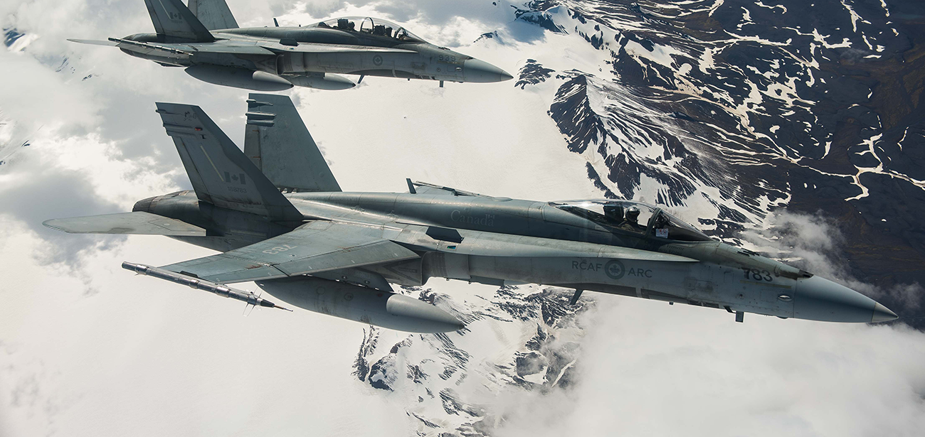 Deux avions de chasse gris de l’Aviation royale canadienne survolent une chaîne de montagnes enneigée.