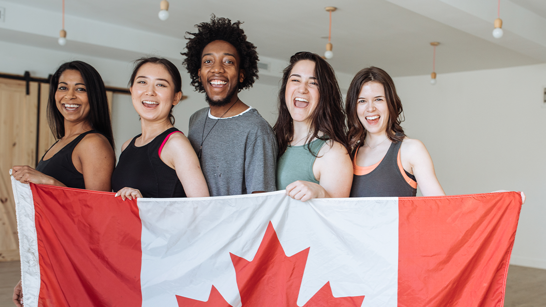 Four young women and one young man stand in a row smiling and holding up a Canada flag. 