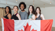 Four young women and one young man stand in a row smiling and holding up a Canada flag. 