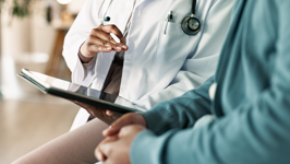 A close-up of a doctor holding a tablet while sitting beside a patient.