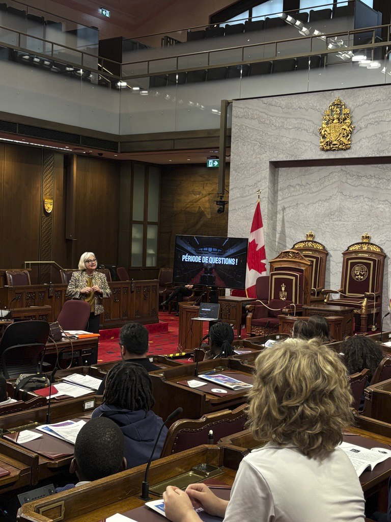 Une femme en blazer s’adresse à des jeunes assis à des pupitres. 