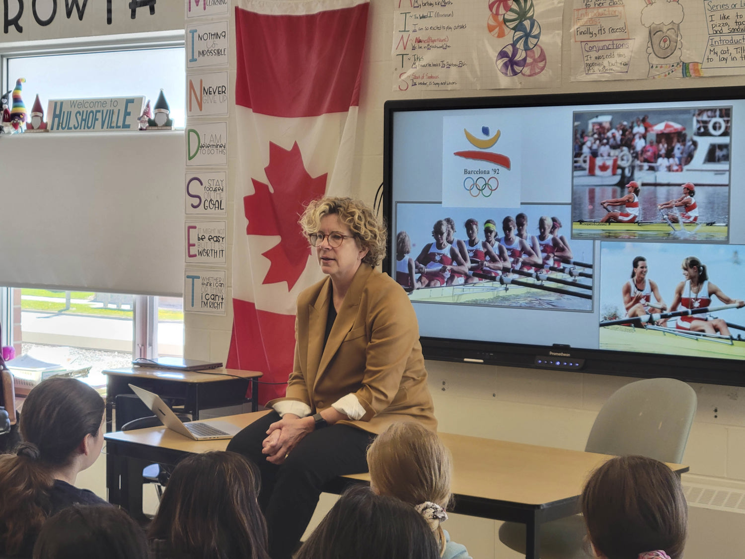 Une femme en blazer, assise sur une table, donne une présentation aux élèves dans une salle de classe.