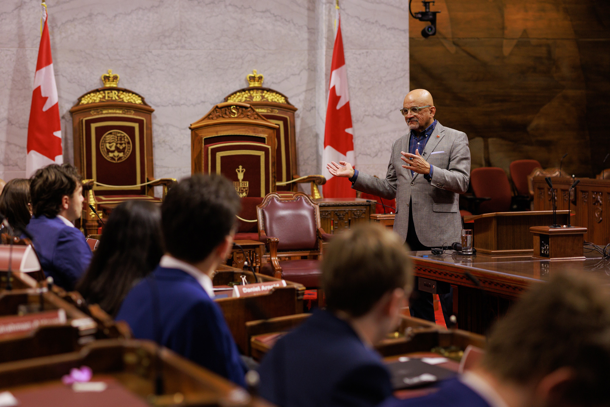 A man speaks to a group of young adults sitting in the Senate Chamber.